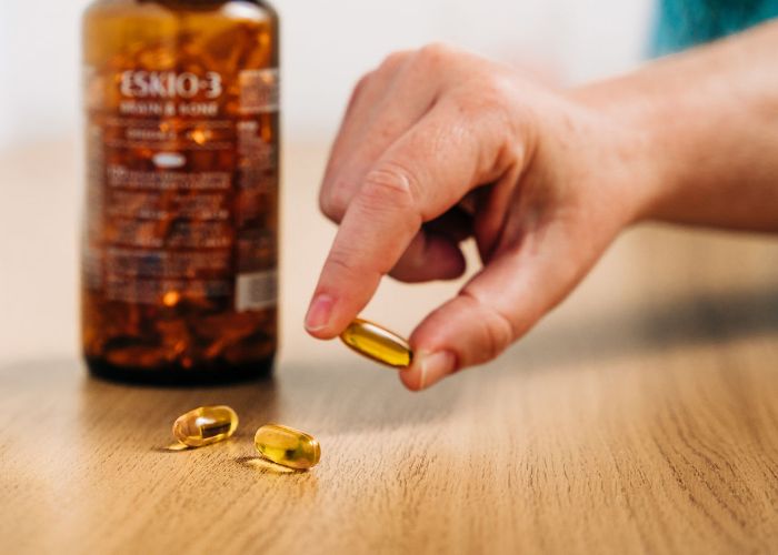 Hand som h&aring;ller en omega-3-kapsel bredvid en burk med fiskolja.